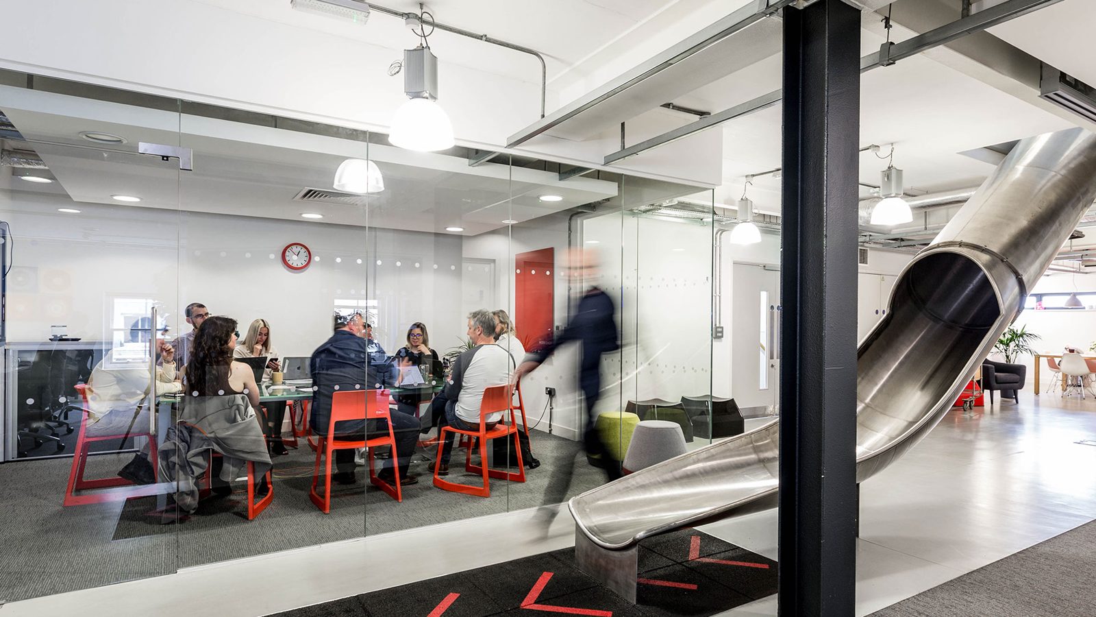 The steel slide between floors, beside a glass-walled meeting room in use